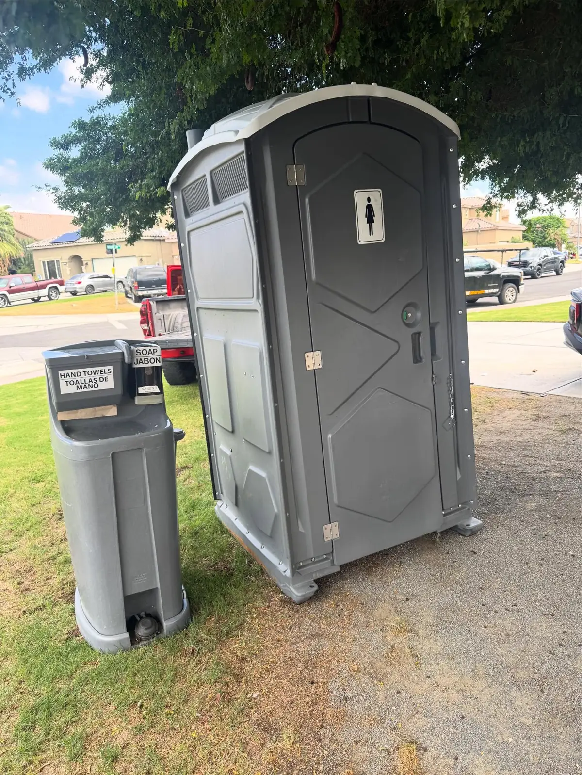 Grey porta potty on a lawn beside a stand-alone hand-wash station